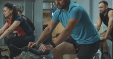 Sakallı adam zamanı spor salonundaki diğer insanlarla birlikte bisiklet sürmek olarak kontrol ediyor. Kendine güvenen beyaz sporcunun egzersiz bisikleti üzerindeki kasları çalıştırışının yan görüntüsü. Yaşam tarzı. Sinema 4k ProRes Merkezi.