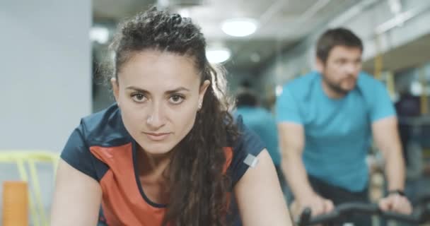 Nahaufnahme Porträt einer brünetten Frau mit braunen Augen, die auf einem Heimtrainer trainiert, während im Hintergrund ein verschwommener Mann reitet. Kaukasische Sportlerin beim Training im Fitnessstudio. Sport, Kraft. Cinema 4k ProRes Hauptquartier. — Stockvideo