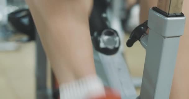 Primer plano de piernas caucásicas masculinas pedaleando bicicleta estática en club deportivo. Deportista deportivo irreconocible en bicicleta en el gimnasio en el equipo de ejercicio. Entrenamiento, ejercicio, fitness. Sede del cine 4k ProRes . — Vídeos de Stock