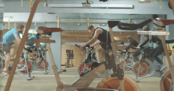 Radosny profesjonalny trener wspierający osoby szybko jeżdżące na rowerach treningowych w siłowni. Szeroki widok z boku sportowca wesoły mężczyzn i kobiet ćwiczących na sprzęt rowerowy. Kino 4k ProRes kwatera główna. — Wideo stockowe