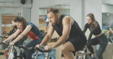 Egzersiz bisikletinde hızlı giden ve egzersizi bitiren yorgun ve terleyen adamın yan görüntüsü. Beyaz tenli yakışıklı esmer sporcu diğer insanlarla birlikte spor salonunda bisiklet ekipmanı eğitimi alıyor. Cinema4k ProResHQ.