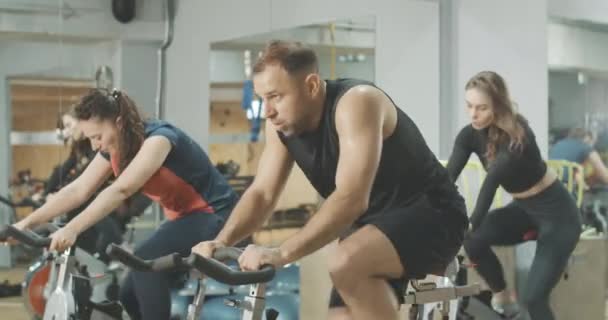 疲れ汗をかく男の側のビューは、運動自転車や運動を終了する上で速く乗っている。他の人とジムでサイクリング機器に関するハンサムな白人のブルネットのスポーツマンのトレーニング。Cinema4k｜ProResHQ. — ストック動画