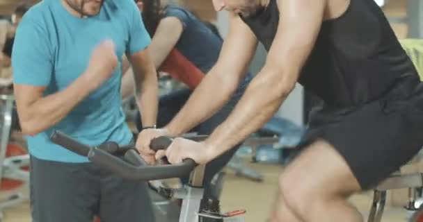 Giovane affascinante allenatore caucasico che sostiene l'uomo traspirante in bicicletta su cyclette in palestra. Allegro dipendente fiducioso che corregge l'allenamento di un forte sportivo. Cinema 4k ProRes HQ . — Video Stock