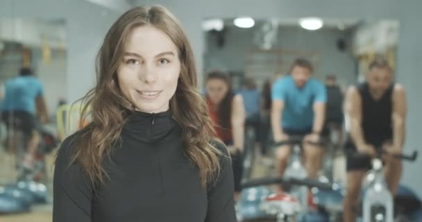 Πορτρέτο της όμορφης γοητευτικής καυκάσιας γυναίκας που ποζάρει στο γυμναστήριο στο παρασκήνιο των ανθρώπων που ποδηλατούν με ποδήλατα γυμναστικής. Χαμογελώντας αυτοπεποίθηση Καυκάσια μελαχρινή κοπέλα με γκρίζα μάτια. Κινηματογράφος 4k ProRes HQ. — Αρχείο Βίντεο