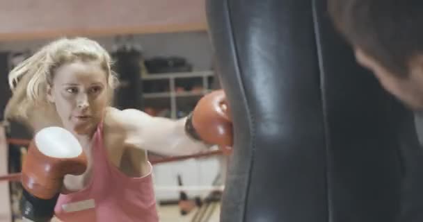 Självsäker blond vit kvinna boxning boxningssäck i sportklubben. Porträtt av koncentrerad kvinnlig boxerträning med tränare på gym. Självförsvar, jämställdhet. Bio 4k ProRes HQ. — Stockvideo