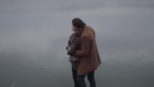 Side view of laughing Caucasian couple hugging at the background of grey water on cloudy day. Positive young man and woman enjoying dating outdoors by lake or river. Love, lifestyle, happiness. — Stock Video