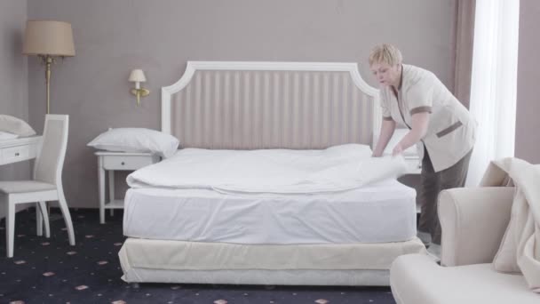 Dospělá běloška ustlala postel v hotelovém pokoji. Široký záběr profesionální zaměstnankyně narovnávající bílou přikrývku. Cestovní ruch, služby, čistota, profesionalita. — Stock video