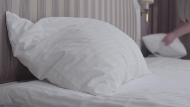 Close-up de travesseiro deitado em grande cama de luxo no quarto de hotel com funcionário borrado colocando almofada na cama. Empregada caucasiana irreconhecível a trabalhar num resort turístico. Trabalho, ocupação, estilo de vida . — Vídeo de Stock