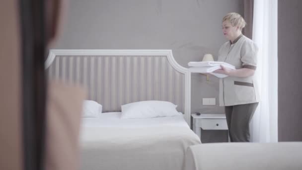 Sidovy av självsäker vit piga sätta rena vita handdukar på sängen i hotellrummet. Professionell arbetare förbereder logi för turister. Livsstil, turism, städning, service. — Stockvideo