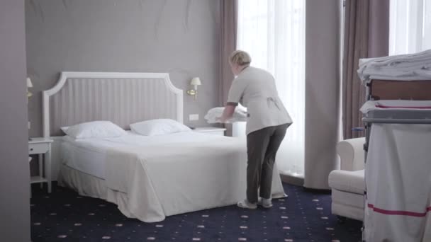 Вид на горничную отеля, готовящую гостиничный номер для посетителей. Широкий снимок профессиональной кавказской женщины, надевающей чистые халаты на большую кровать в спальне на курорте. Мбаппе, сервис, стиль жизни . — стоковое видео