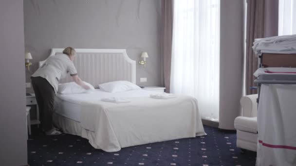 Szerokie ujęcie pokoju hotelowego z białą pokojówką ścielącą łóżko. Profesjonalna wykwalifikowana kobieta dostosowuje poduszki i koc prostowania w zakwaterowaniu. Serwis, turystyka, kurort. — Wideo stockowe