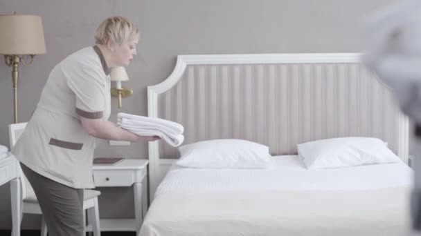 Mellanslag av allvarliga piga sätta rena handdukar på vit säng och lämnar. Sidovy porträtt av professionell vit kvinna i uniform gör städning i hotellrum. Turism, sysselsättning, tjänster. — Stockvideo