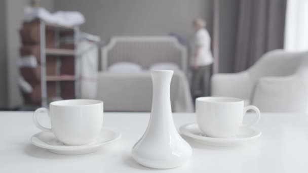 在阳光下，茶杯和茶壶的特写站在桌子上，作为模糊的女佣擦去床上的灰尘。专业的白人女性清洁酒店房间。旅游、服务、工作. — 图库视频影像