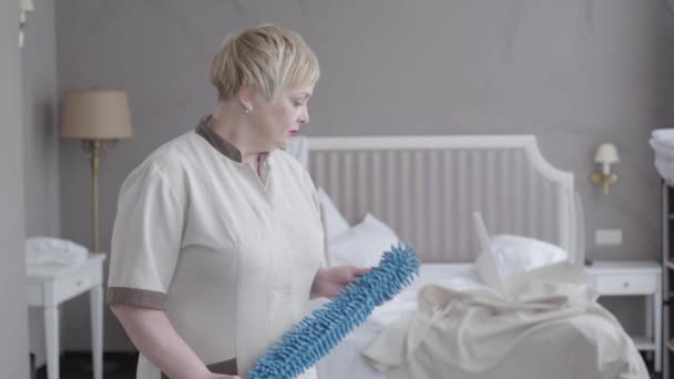 Exhausted professional maid looking back at bed in hotel room and turning to camera. Middle shot portrait of Caucasian adult woman in uniform posing with dust brush. Job, occupation, service. — Stock Video