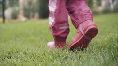 Kamera, yeşil ıslak çimlerde gezinen pembe çizmeli çocuğun bacaklarını takip ediyor. Yağmurlu bir günde dışarıda yürüyen tanınmayan bir kız. Boş zaman, çocukluk, yaşam tarzı.