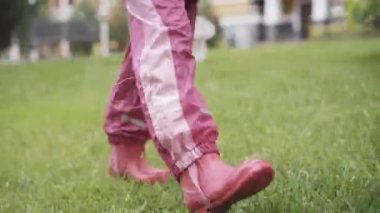 Pembe lastik çizmeli ve su geçirmez pantolonlu küçük bir kız ıslak yeşil çimlerde yürüyor. Yağmurdan sonra dışarıda gezinen kendinden emin bir çocuk. Boş zaman, eğlence, yaşam tarzı, çocukluk, eğlence