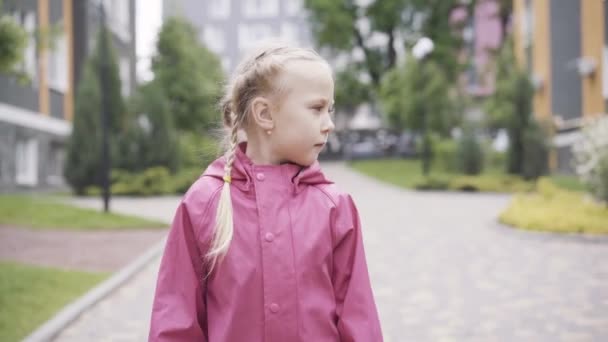 Retrato de una linda niña con abrigo impermeable caminando por el callejón al aire libre. Niño bastante caucásico en ropa rosa paseando en el día lluvioso. Infancia, ocio, estilo de vida . — Vídeos de Stock