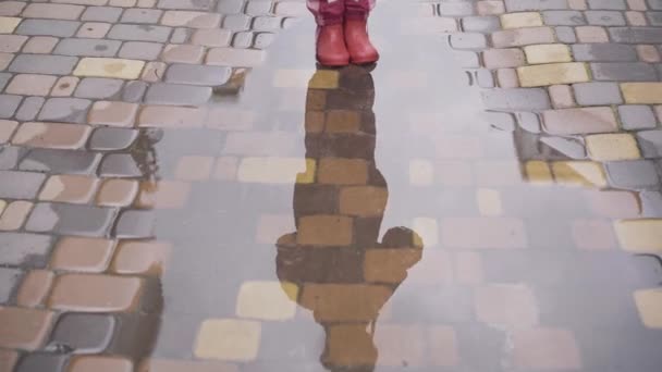 Silueta de niña reflejándose en charco sobre adoquines. Niño irreconocible con botas de goma rosa al aire libre después de la lluvia. Temporada de lluvias, clima húmedo, infancia, estilo de vida . — Vídeos de Stock