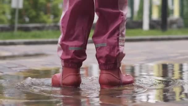 Вид сзади на неузнаваемую девочку, стоящую в луже. Ребенок в розовых штанах и красных резиновых сапогах наслаждается дождливым днем на открытом воздухе. Детство, развлечения, радость, досуг, образ жизни . — стоковое видео