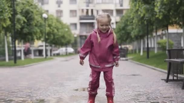 Retrato de niña alegre divirtiéndose jugando en charco en el día lluvioso. Amplio plano de alegre niño rubio caucásico descansando al aire libre. Diversión, alegría, ocio, infancia . — Vídeos de Stock