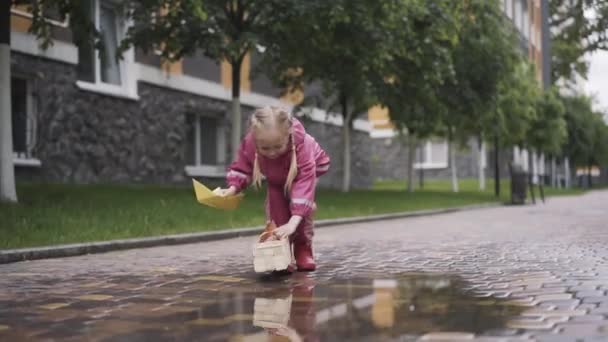 Glückliches Mädchen in pinkfarbener Kleidung, das neben einer Pfütze mit Papierbooten spielt. Weitwinkelporträt eines positiven kleinen kaukasischen Kindes, das einen regnerischen Tag im Freien genießt. Freizeit, Freude, Kindheit, Lebensstil. — Stockvideo