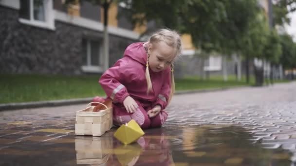 Jolie petite fille soufflant sur le bateau en papier jaune pour qu'il coule sur la flaque d'eau. Portrait d'enfant caucasien joyeux assis sur le trottoir et jouant avec le jouet. Enfance, loisirs, joie, plaisir . — Video