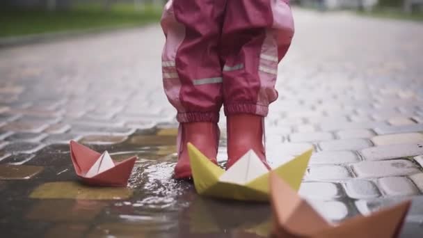 ゴム製のブーツを着た少女の足が紙のボートで水たまりの中を歩いている。雨の日に屋外で遊ぶことを楽しんでいる認識できない幸せな子供。子供時代、レジャー、楽しみ. — ストック動画