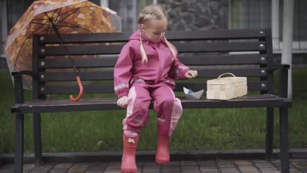 Menina caucasiana alegre sentado no banco e brincando com barco de papel. Grande tiro de criança pequena feliz descansando ao ar livre no dia chuvoso com brinquedo. Alegria, lazer, infância, estilo de vida . — Vídeo de Stock