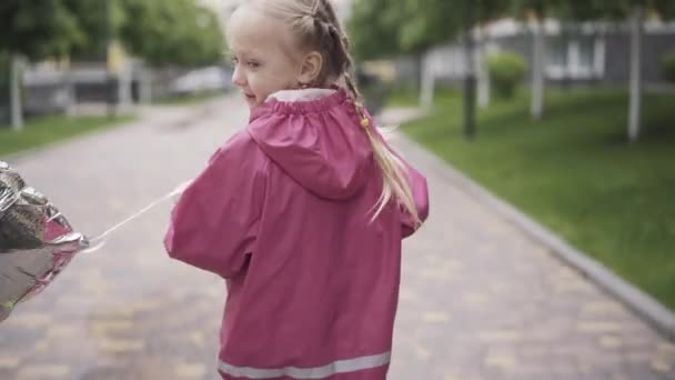 風船に乗ってスクーターと陽気なブロンドの女の子のバックビュー。雨の日に屋外で週末を楽しむ白人の子供の肖像画。カメラは顔から足に下に移動します。子供時代、レジャー. — ストック動画