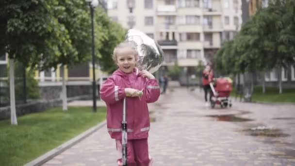 Retrato de alegre chica feliz montando scooter al aire libre. Riendo lindo niño caucásico sosteniendo globo y divertirse en el día lluvioso. Infancia, ocio, alegría . — Vídeos de Stock