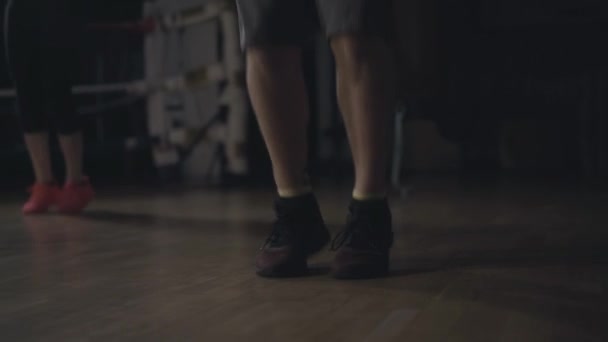 Primer plano de los pies caucásicos masculinos saltando la cuerda en el gimnasio oscuro. Mujer joven irreconocible haciendo ejercicio en el fondo. Fitness, deporte, estilo de vida, entrenamiento . — Vídeos de Stock