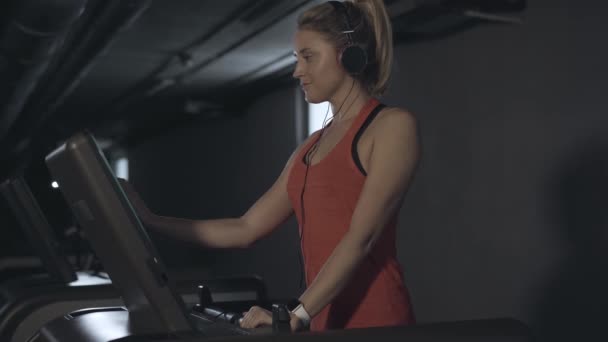 Vista lateral de la joven rubia sonriente ajustando los ajustes en la cinta en el gimnasio. Retrato de la deportista caucásica feliz en el entrenamiento de auriculares en el club de fitness. Deporte, salud, estilo de vida . — Vídeo de stock