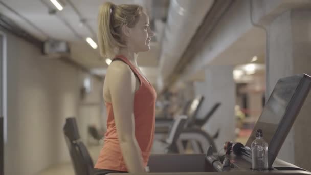 Retrato de vista lateral de una hermosa mujer caucásica confiada que apaga la cinta y se va. Joven rubia atleta caucásica terminando el ejercicio en el gimnasio. Deporte, fitness, estilo de vida . — Vídeo de stock