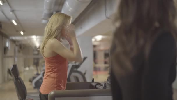 一个积极的金发女运动员在跑步机上行走，与一位无法辨认的高加索女性朋友交谈。年轻迷人的女运动员在体育馆训练的侧视图。体育、运动、生活方式. — 图库视频影像