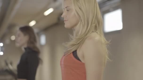 Prachtige zelfverzekerde sportvrouw die op de loopband loopt en naar de camera draait. Portret van charmante blanke vrouw training in de sportschool met wazig brunette vrouwelijke vriend oefenen op de achtergrond. — Stockvideo