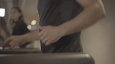 Kamera, erkek bacağından yüze, koşu bandında egzersiz yapan ve arka planda genç zayıf kadın eğitimiyle konuşan sporcunun pozitifliğini gösteriyor. Spor, spor, sağlıklı yaşam tarzı, sağlık..