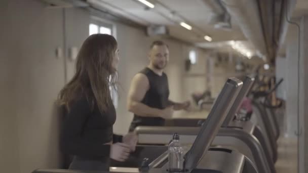 Πλευρική άποψη της καυκάσιας γυναίκας που κάνει τζόκινγκ στο διάδρομο και μιλάει με τον θολή όμορφο Καυκάσιο άντρα που τρέχει στο βάθος. Άσκηση των αθλητικών ανθρώπων στο γυμναστήριο. Υγιεινός τρόπος ζωής. — Αρχείο Βίντεο