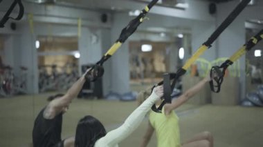 Güçlü kendine güvenen atletik insanlar spor salonunda TRX kayışlarıyla çalışıyor. Genç beyaz erkek ve kadınlar ellerinde ekipman ve egzersiz tutuyorlar. Spor, sağlık, dayanıklılık, eğitim..