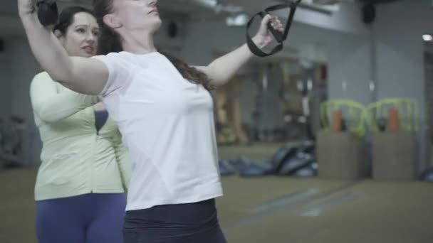 Profesionální trenérka trénuje mladou brunetku v tělocvičně pomocí TRX popruhů. Portrét sebevědomé soustředěné bělošské sportovkyně cvičící s osobním trenérem. Wellness, zdravý životní styl. — Stock video
