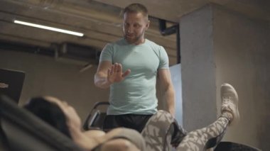 Pozitif gülümseyen adam yer çekimi egzersizi yapan sporcu kadını eğitiyor. Kendine güvenen, beyaz tenli antrenör, bacağı havada, çömelmiş atletik bir kadınla konuşuyor. Spor, egzersiz, sağlık..