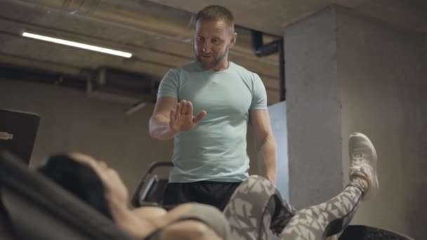 Pozitivně se usmívající muž trénuje sportovkyni při gravitačním cvičení. Sebevědomý dospělý bělošský trenér mluví s atletickou ženou dřepící s nohama nad hlavou. Sport, cvičení, wellness. — Stock video