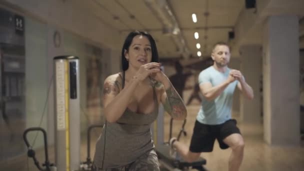 Atractiva entrenadora personal femenina mostrando ejercicio al hombre borroso repitiendo en el fondo. Retrato de mujer blanca hermosa y confiada con manos tatuadas entrenando deportista en el gimnasio . — Vídeos de Stock