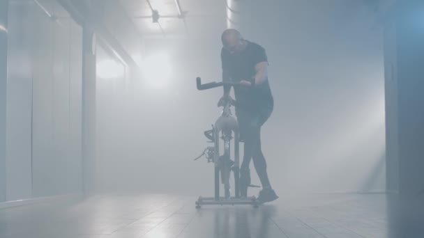 在雾蒙蒙的健身房里，许多运动员骑着健身自行车开始骑马。一个自信的白种人秃头男子在迷蒙的健身俱乐部里锻炼的画像。健康、体育、生活方式、培训. — 图库视频影像