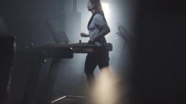 Sisli spor salonunda arka planda koşan sportif bir kadın. Esmer fitness kulübünde koşu bandında koşan beyaz bir kadının geniş açılı görüntüsü. Sağlıklı yaşam tarzı, eğitim, spor, güven.