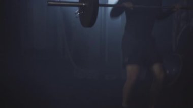 Geniş açılı, yakışıklı esmer sporcu sisli arka planda ağırlık kaldırıyor. Kaslarını sisli spor salonunda çalıştıran güçlü, kendine güvenen beyaz adamın portresi. Vücut geliştirme, spor, yaşam tarzı.