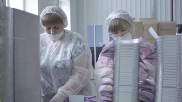 Dos trabajadores adultos de la fábrica caucásica en máscaras faciales y uniformes hablando y empacando envases de alimentos vacíos en la fábrica. Retrato de mujeres que trabajan en la línea de producción durante la cuarentena pandémica de Covid-19 . — Vídeo de stock