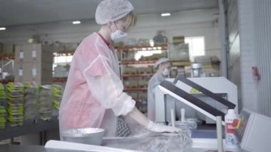 Yüzü maskeli, üniformalı, süpürge makinesiyle yiyecek paketleyen kendine güvenen beyaz bir kadın. Covid-19 karantinasındaki üretim hattındaki profesyonel fabrika işçisinin portresi.