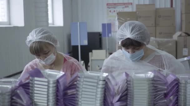 Portret dwóch dorosłych białych kobiet w maskach pracujących w fabryce. Blondynka i brunetka pakują pojemniki na jedzenie na linii produkcyjnej. Produkcja, zatrudnienie, pandemia Covid-19 — Wideo stockowe