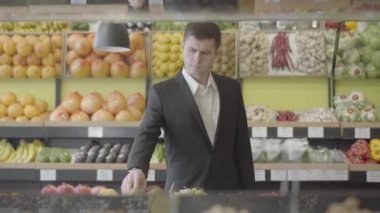 Takım elbiseli ciddi bir beyaz adam markette kivi meyvesi seçiyor. Kendine güvenen yetişkin işadamının portresi perakende dükkanından çilek alıyor. Tüketim, vejetaryenlik, sağlıklı beslenme, vitamin gıdası. S-log 2.