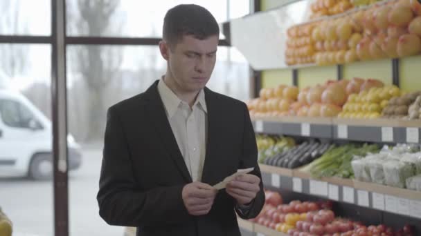 Porträtt av rika kaukasiska affärsman undersöka priset på mat i proposition och suckande. Ung man i kostym gör inköp i matvaror. Livsstil, konsumism, shopping. S-log 2. — Stockvideo
